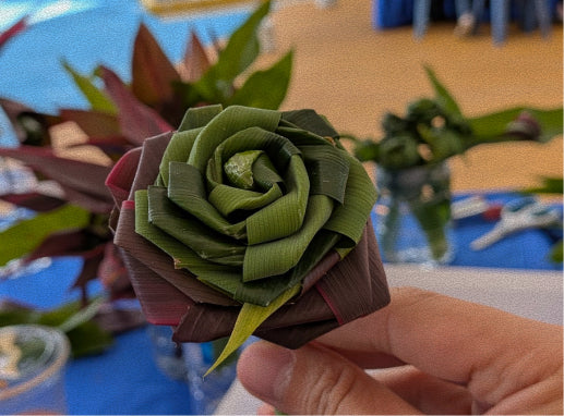 A hand holding a handmade green leaf rose decoration, with a blurred workshop or garden background containing other greenery and blue surfaces.