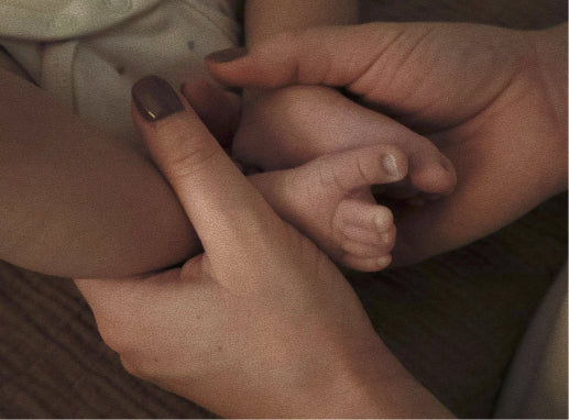 A close-up of an adult's hands holding and cradling a baby's hand and foot, with the adult's nails painted in a dark metallic brown shade.