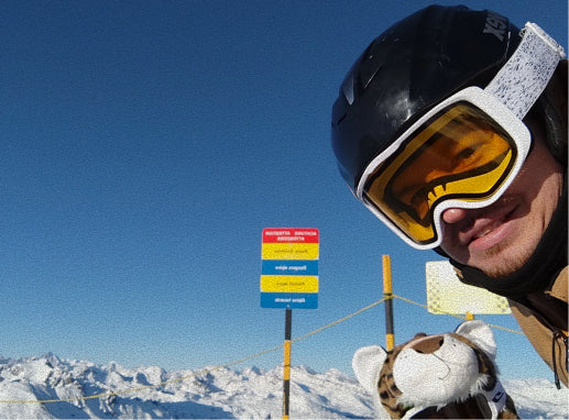 A close-up selfie of a person wearing a black ski helmet and white-framed goggles with yellow lenses, standing on a snowy mountain under a clear blue sky. A small stuffed animal is visible at the bottom, and a ski resort sign is in the background.
